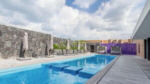 Piscina com degraus de acesso, rodeada por espreguiçadeiras e guarda-sóis, com uma parede de pedra e um edifício moderno ao fundo.