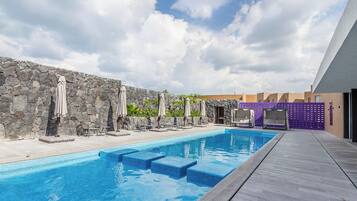 A swimming pool with steps leading into it, surrounded by lounge chairs and umbrellas, with a stone wall and a modern building in the background.