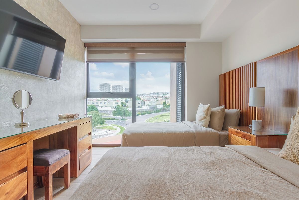 A modern hotel room with a large bed, a desk with a mirror, a television, and a view of the cityscape.