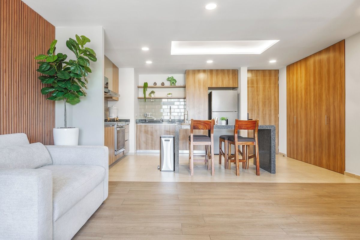 Cozinha moderna com armários de madeira, área de jantar com mesa e cadeiras, e sala de estar com sofá e planta em vaso.