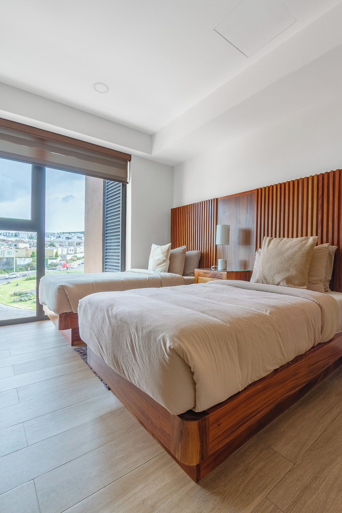 A modern bedroom with a large bed, wooden headboard, and a view of a residential area through a sliding glass door.