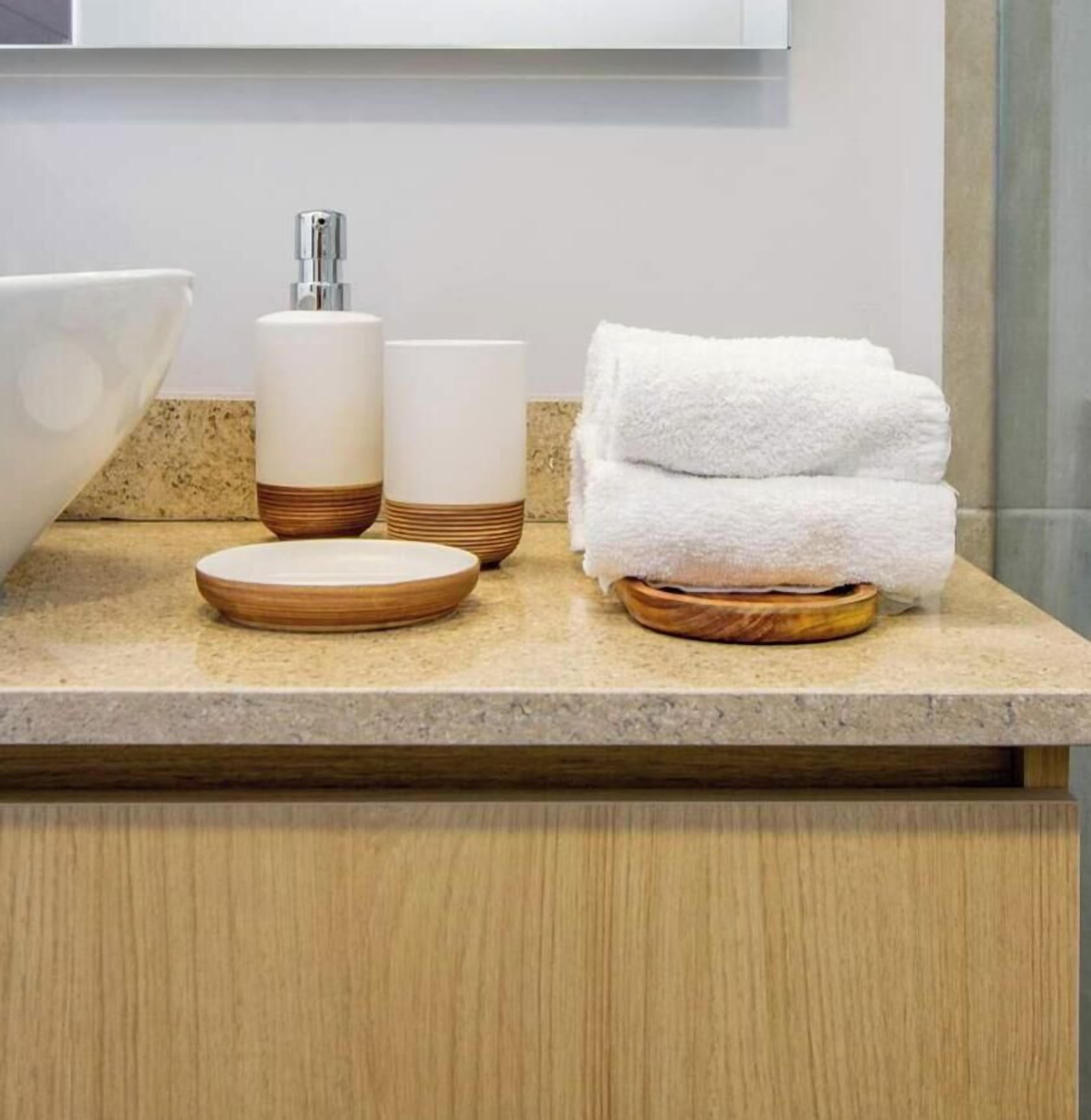 A bathroom vanity with a white sink, a bottle of liquid soap, a cup, and a stack of white towels.