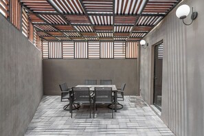 Een overdekt terras met een tafel en stoelen, een plafond met houten latten en een wandlamp.