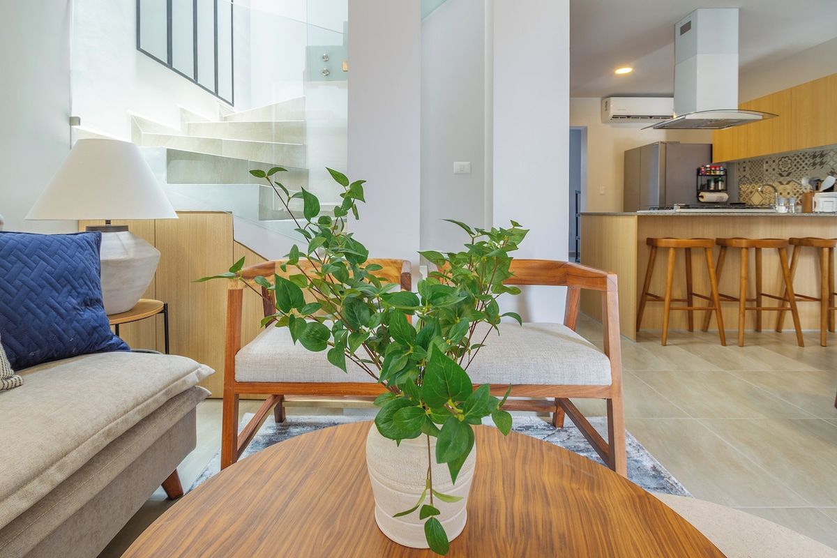 En moderne stue med sofa, sofabord og en plante. Der er et køkkenområde med en barstol og en trappe i baggrunden.
