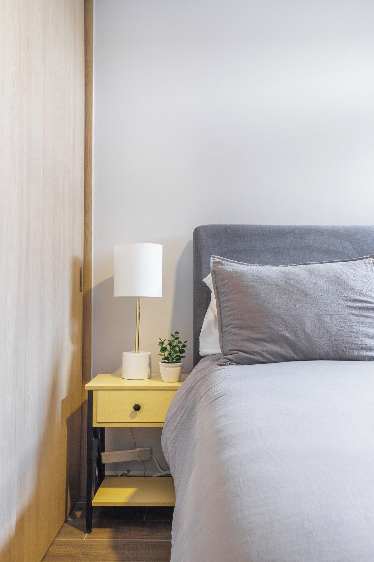 A neatly made bed with a grey headboard, a bedside table with a lamp, and a small potted plant.