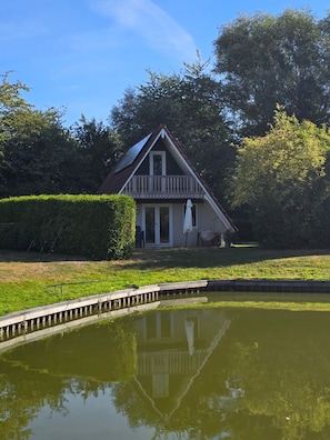A house with a balcony, a pond in front, and a hedge to the left. - Luxury pond lodge with a wood-burning stove on a fishing lake (Gramsbergen)