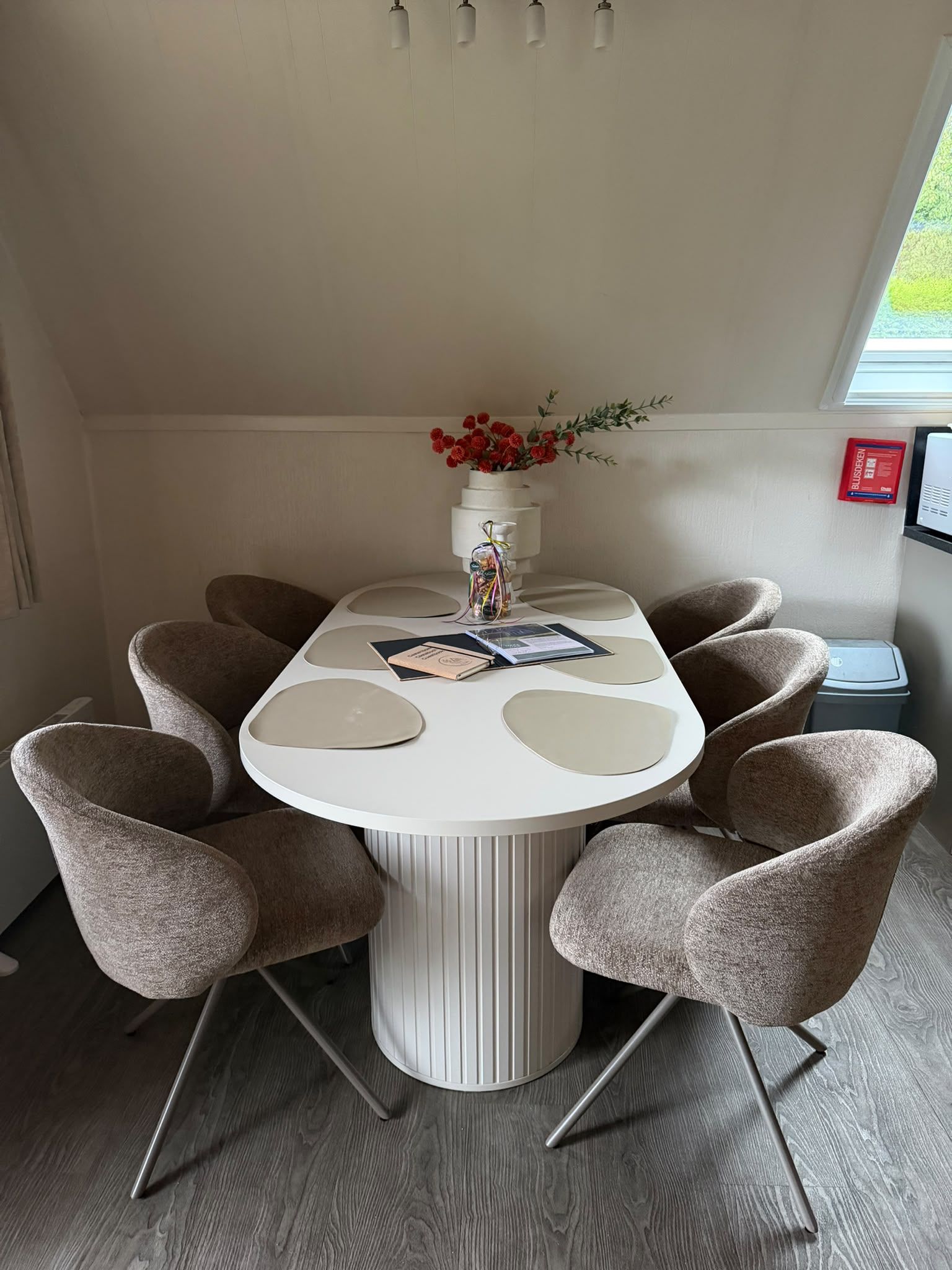 Un comedor con una mesa redonda blanca rodeada de seis sillas, un florero con flores y una ventana con persianas.