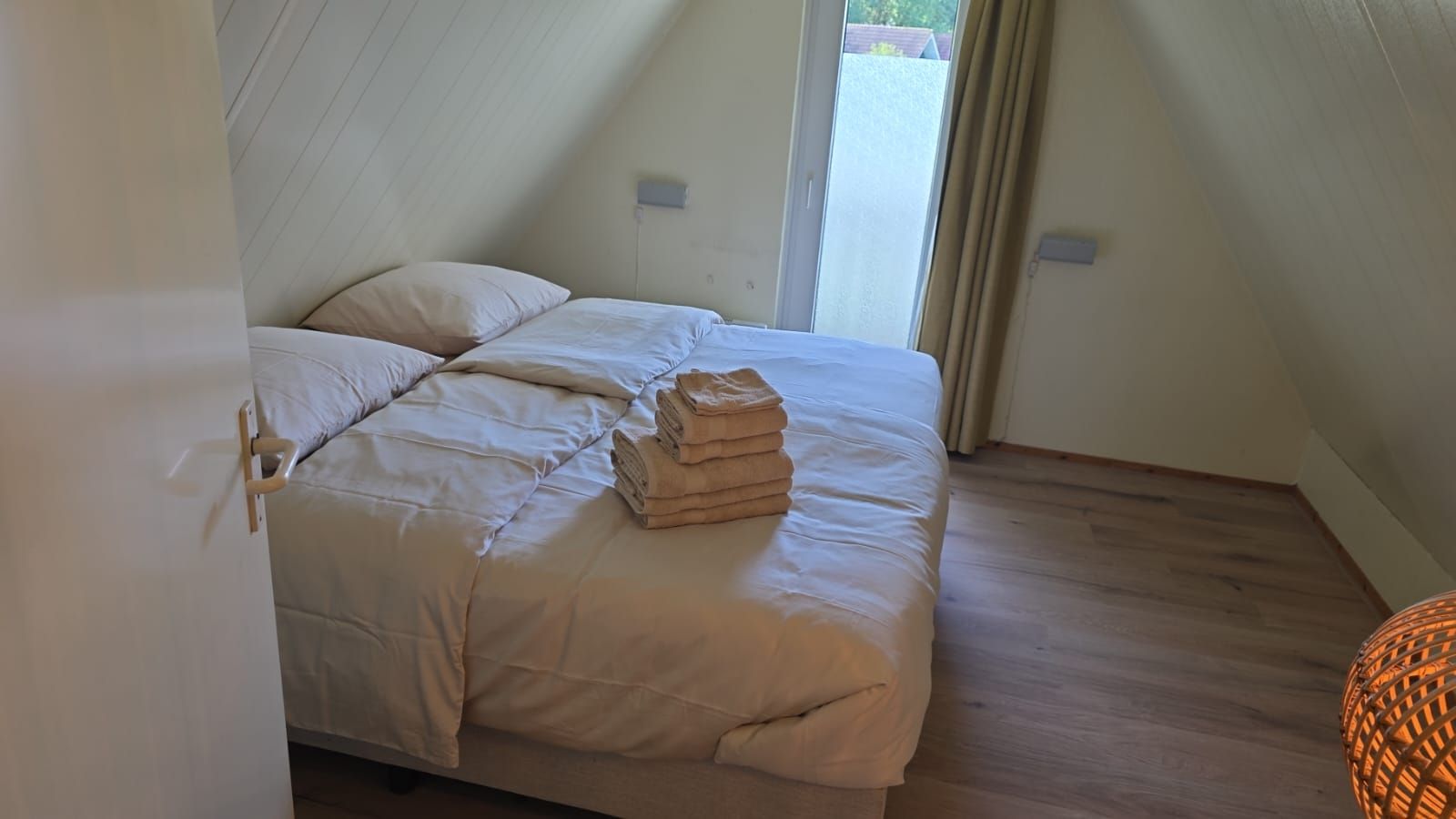 Ein Einzelbett mit einer beigen Bettdecke und Kissen, ein Stapel gefalteter Handtücher und ein Fenster mit Vorhängen.
