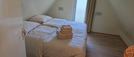 Un letto singolo con copriletto e cuscini color beige, una pila di asciugamani piegati e una finestra con tende.