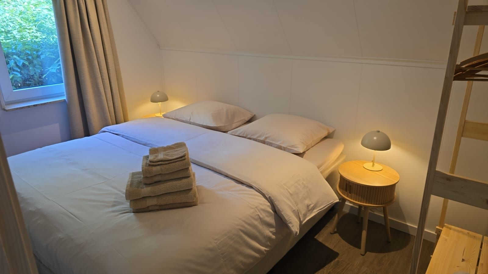 A bedroom with two beds, a window with curtains, a bedside lamp, and a stack of towels.