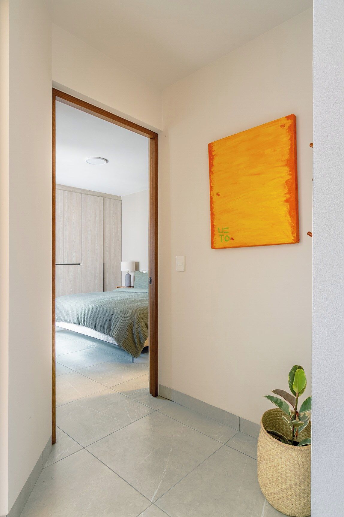 Corredor que dá para um quarto com cama, um quadro na parede e um vaso com planta.