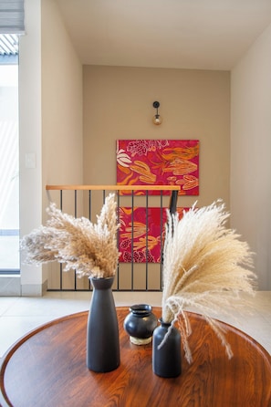Une pièce avec un plancher en bois, une table ronde en bois ornée de deux vases et de quelques plumes d’herbe des pampas, un luminaire fixé au mur et un tableau abstrait rouge.