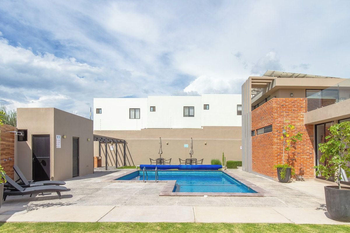 A swimming pool with lounge chairs, a covered seating area, and a building with a flat roof and brick facade.