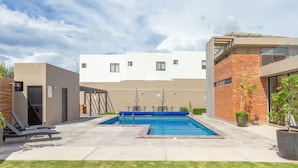 Ein Schwimmbecken mit Liegestühlen, einer überdachten Lounge und einem Gebäude mit flachem Dach und Backsteinfassade.
