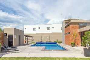 A swimming pool with lounge chairs, a covered seating area, and a building with a flat roof and brick facade. - Casa Colorin | Cosmos Homes (Zakia)
