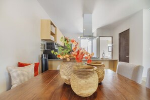 A dining area with a wooden table, three ceramic vases with flowers, and a kitchen in the background. - Casa Colorin | Cosmos Homes (Zakia)