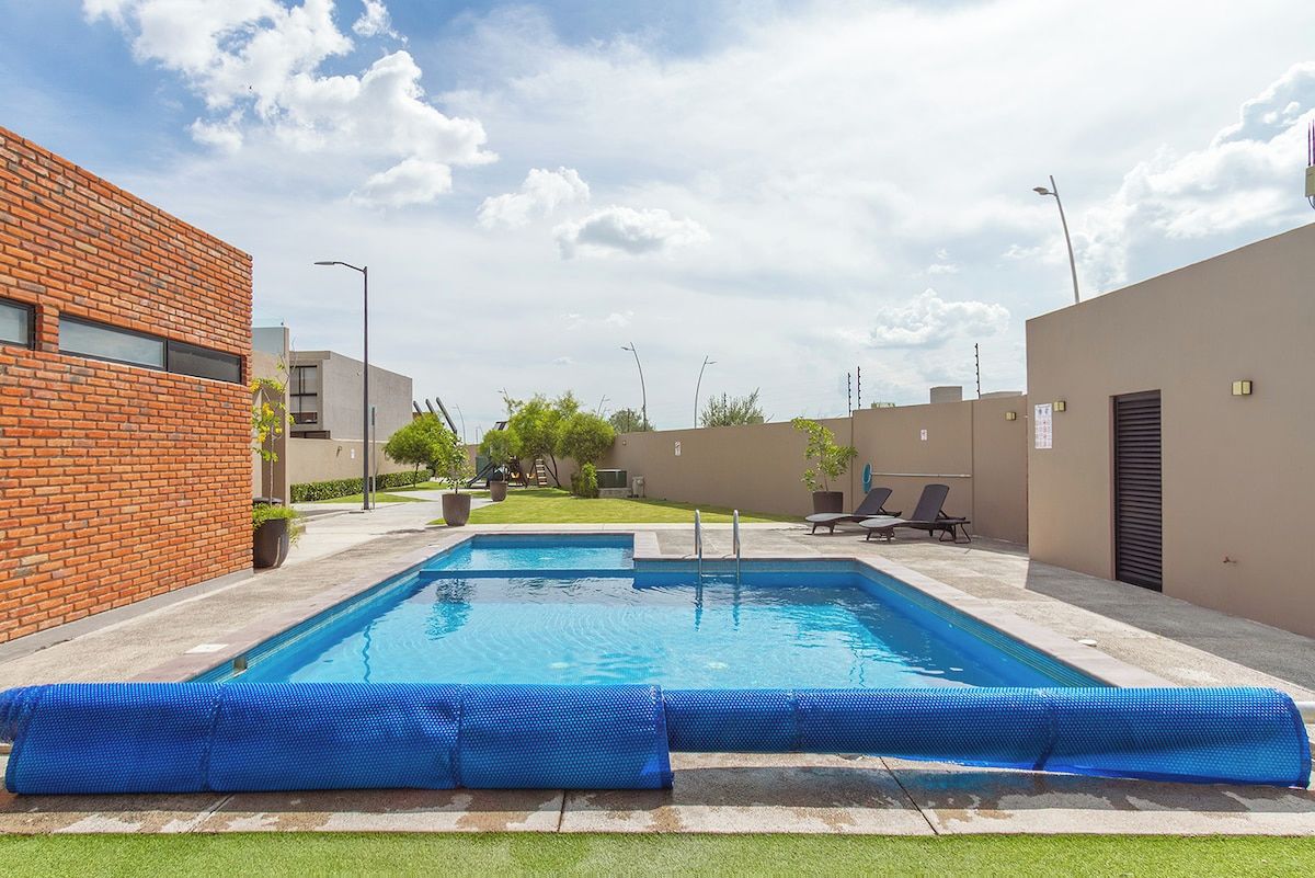 A rectangular swimming pool with a blue border, surrounded by a paved area with a brick building on one side and a plain wall on the other.