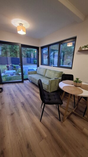 Ein Zimmer mit Holzboden, einem grünen Sofa, einem schwarzen Sessel, einem kleinen Tisch mit einer gewebten Unterlage und einem großen Fenster mit Blick ins Freie.