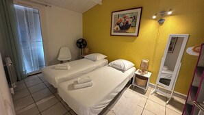 Ett hotellrum med två sängar, en bordsfläkt, en sänglampa, en spegel och ett fönster med gardiner.
