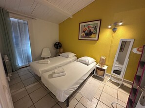 Ein Hotelzimmer mit zwei Betten, einem Standventilator, einer Nachttischlampe, einem Spiegel und einem Fenster mit Vorhängen.