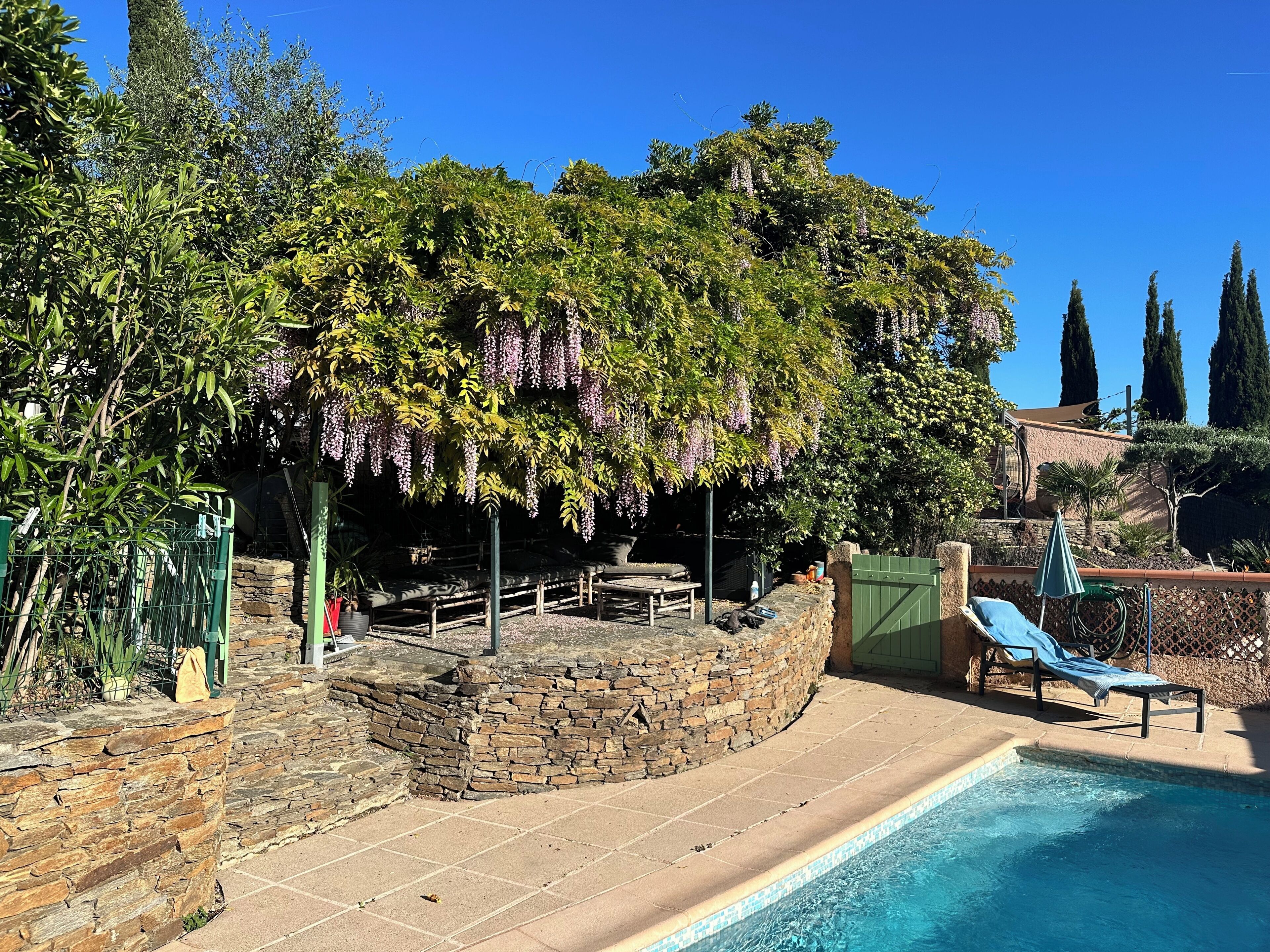 Zona de piscina con muros de piedra, una tumbona y una pérgola con plantas colgantes.