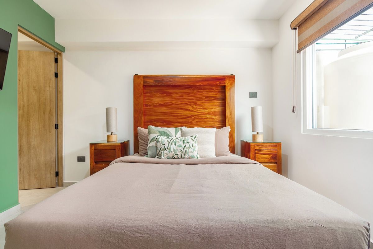 A bedroom with a large bed, wooden headboard, two bedside tables with lamps, a green wall, and a window with blinds.