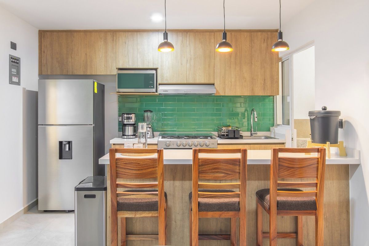 A compact kitchen with wooden cabinets, a stainless steel refrigerator, a microwave, a stove, and a sink. There are three wooden chairs with cushioned seats.
