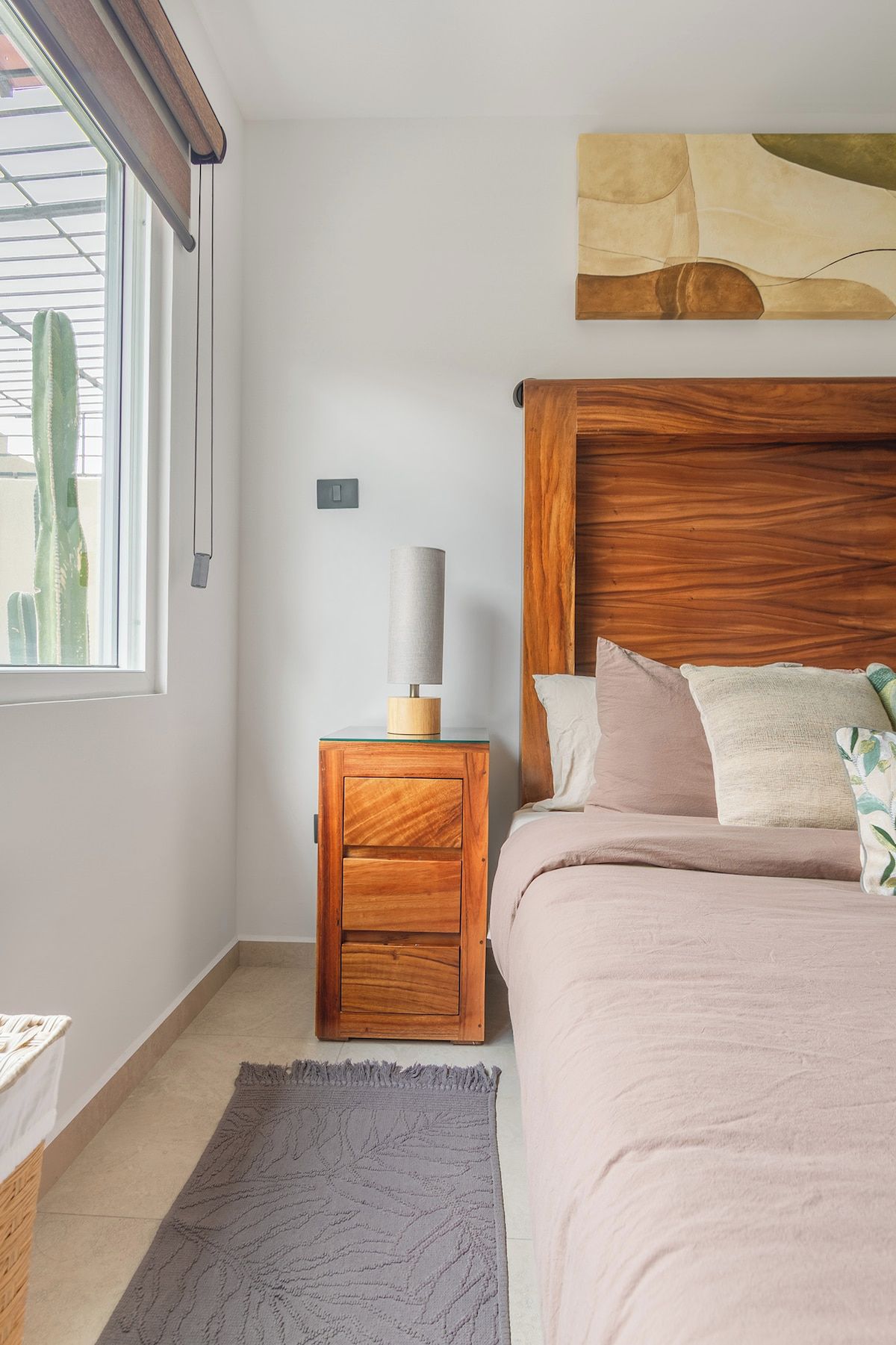 Quarto com cabeceira de madeira, mesa de cabeceira com luminária, cama com travesseiros, janela com vista para um cacto e tapete no chão.