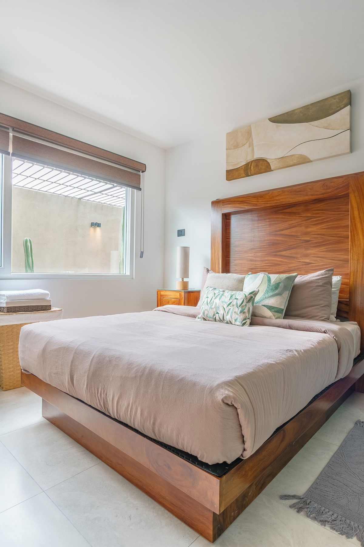 A modern bedroom with a large bed, a bedside table, a nightstand, a lamp, and a window with blinds.