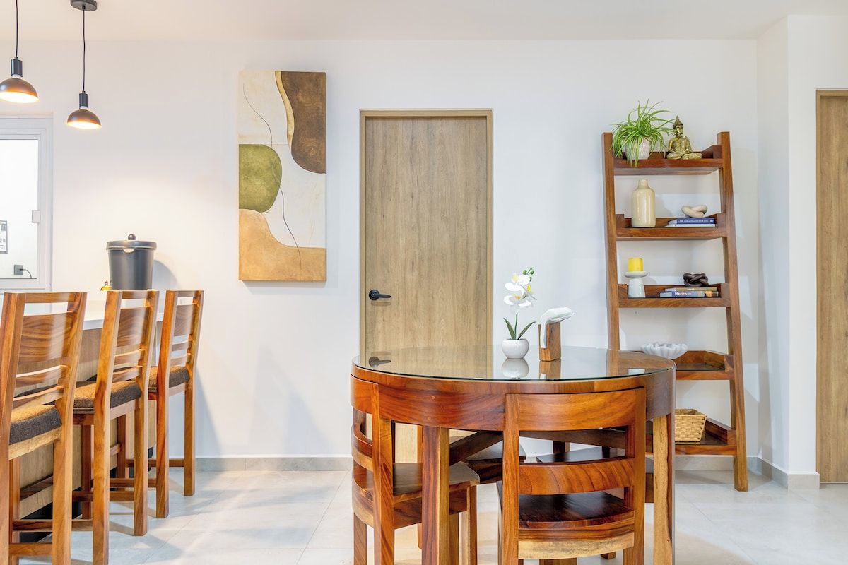 A dining area with a round glass table and wooden chairs, a wooden door, a shelf with decorative items, and pendant lighting.