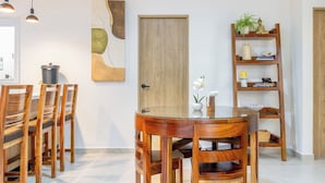 Zona pranzo con tavolo rotondo in vetro e sedie in legno, una porta in legno, una mensola con oggetti decorativi e illuminazione a sospensione.