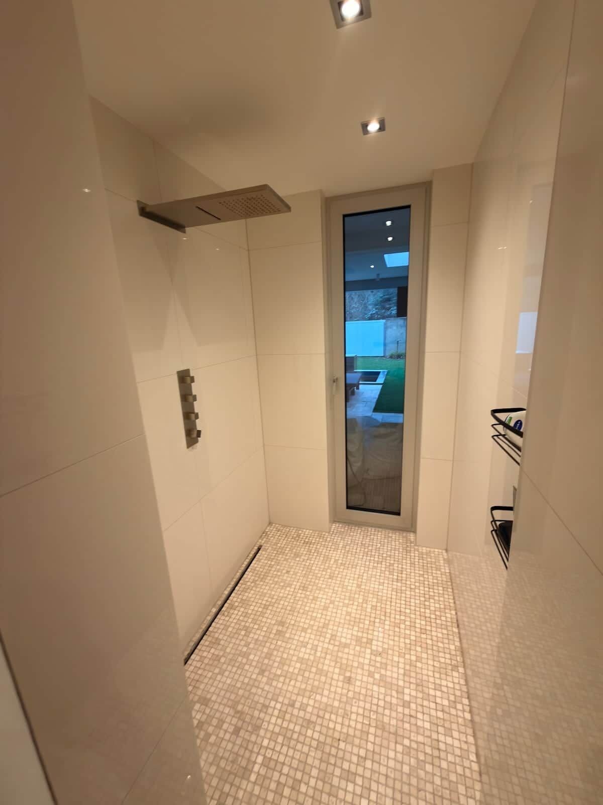 Ein modernes Badezimmer mit Fliesenboden, einer Duschkabine und einer Glastür zu einem Außenbereich.