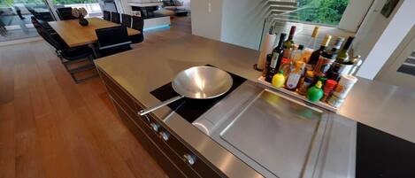 A modern kitchen with a stainless steel stove, a dining area with a wooden table and chairs, and a view of greenery outside.