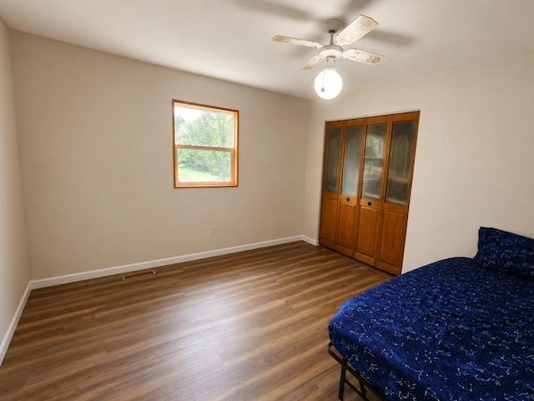 A bedroom with a wooden floor, a bed with a blue cover, a wardrobe with glass doors, a ceiling fan, and a window.