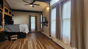 Quarto com piso de madeira, cama com manta listrada, ventilador de teto e janelas amplas com cortinas.