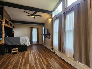 A room with a wooden floor, a bed with a striped blanket, a ceiling fan, and large windows with curtains. - Tranquil 3-bedroom Mountain View Farmhouse in Charming Greeneville (Greeneville)