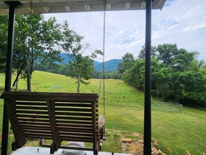 A wooden swing chair under a covered area with a view of a grassy field and trees.