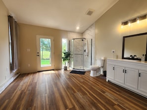 A spacious bathroom with a wooden floor, a glass shower enclosure, a white toilet, and a vanity with a mirror. - Tranquil 3-bedroom Mountain View Farmhouse in Charming Greeneville (Greeneville)