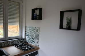 Una cocina con estufa a gas, una ventana con persianas, un estante en la pared con una lata y un espejo enmarcado.