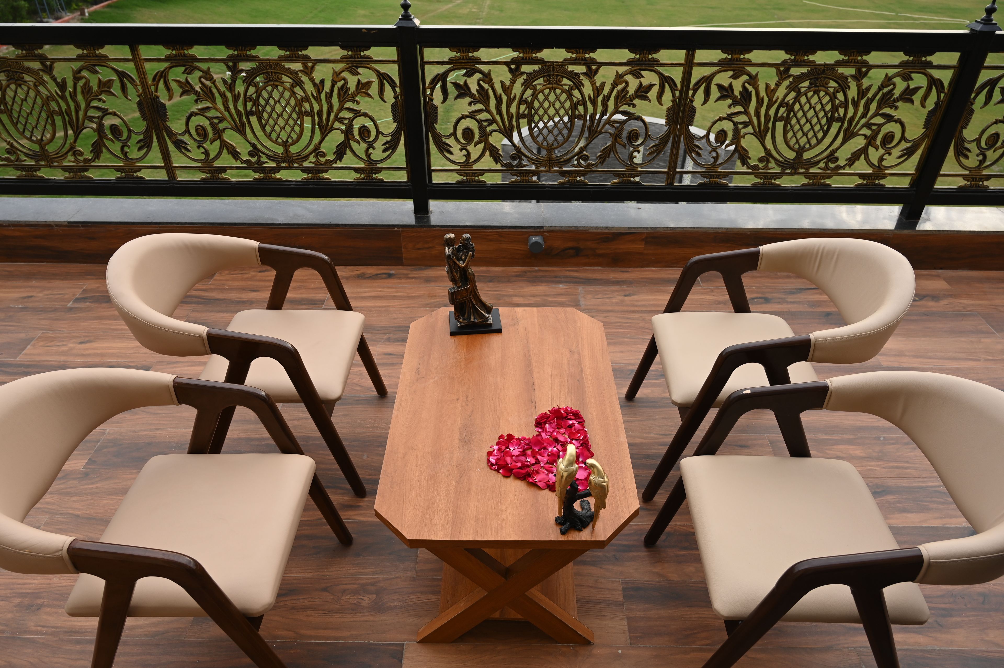 Una mesa de madera con cuatro sillas, una estatua y un arreglo de flores rojas en forma de corazón en un balcón con barandilla decorativa.