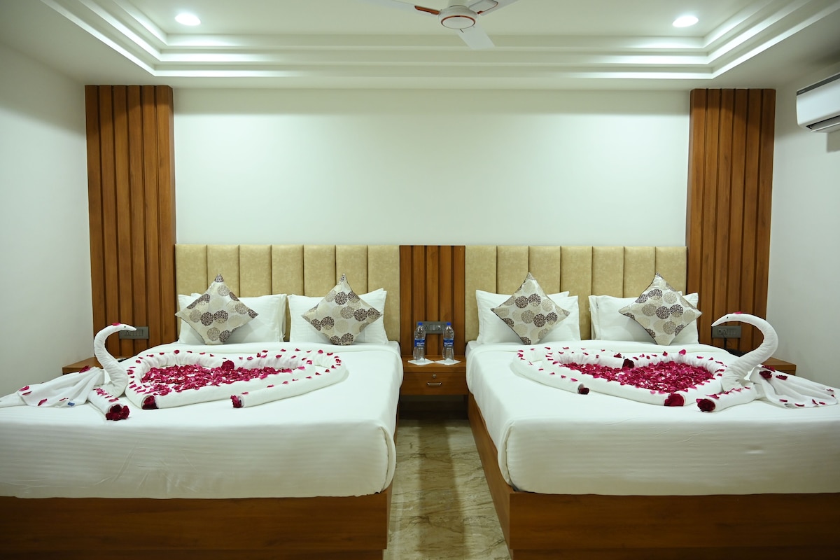 A hotel room with two beds, each with a swan-shaped towel and rose petals. There are decorative pillows, a nightstand with water bottles, and a ceiling fan.