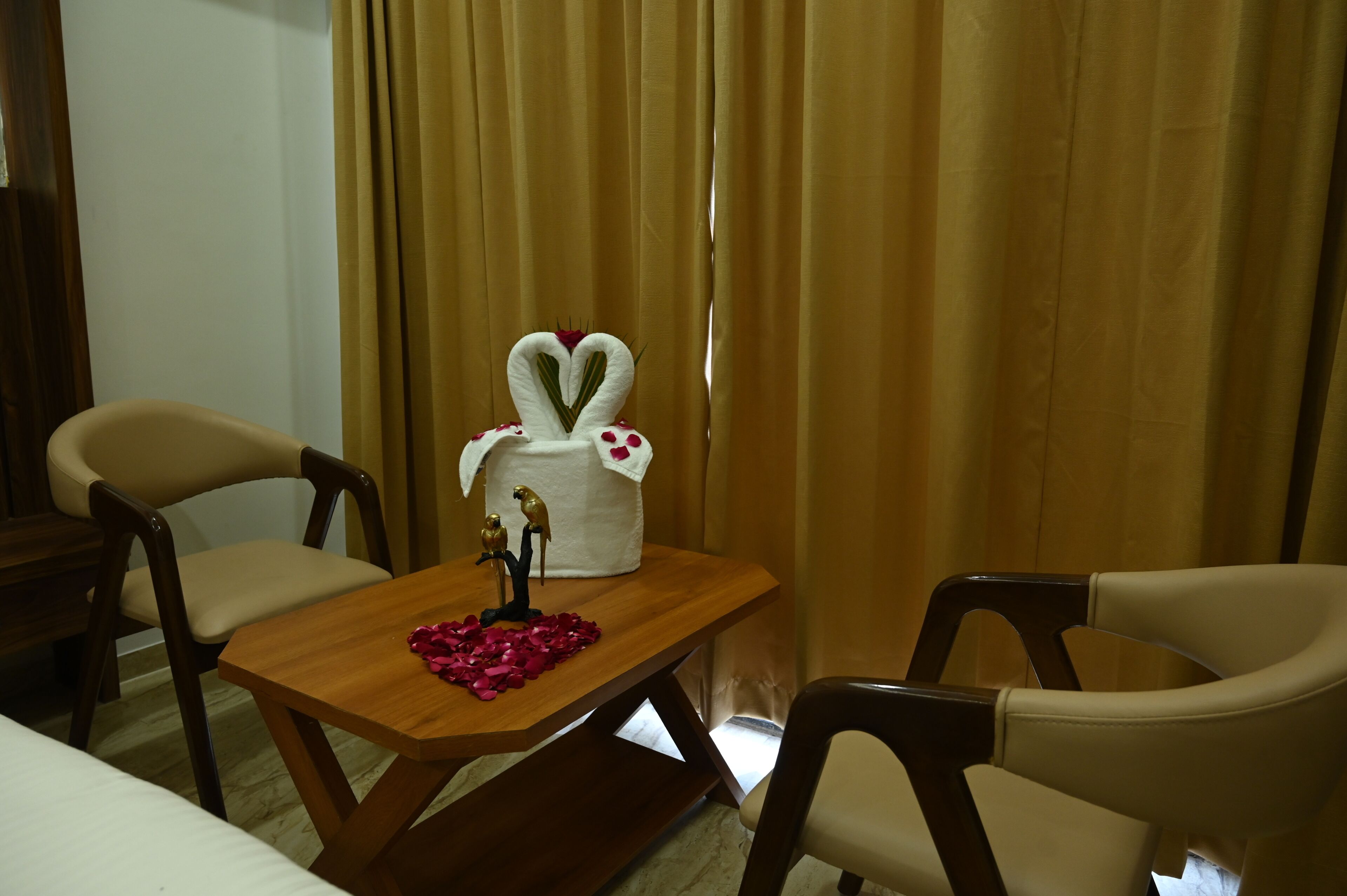 A room with a wooden table, two chairs, a decorative centerpiece with a heart-shaped towel and a parrot figurine, and a curtain in the background.