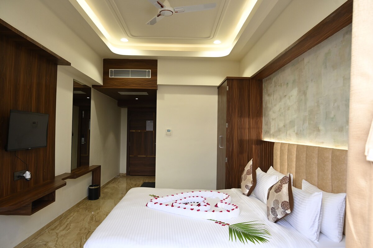 A hotel room with a bed, a ceiling fan, a mounted TV, a wooden wardrobe, and a small trash bin.