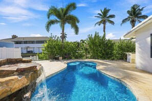 En pool med en böjd kant, omgiven av en stenbelagd terrass, palmer och en vit byggnad.