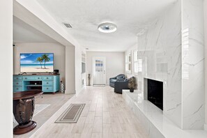 A spacious living room with a fireplace, a television, and a view of the beach through a window.