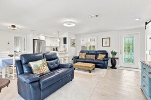 A modern living room with a blue leather sofa, a dining area, and a kitchen with white cabinets.