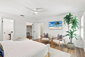 Une chambre à coucher avec un lit, une chaise, une table, une plante et un tableau représentant une plage.
