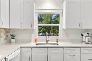 Une cuisine avec des meubles blancs, un évier en acier inoxydable, une fenêtre donnant sur des palmiers, une machine à café et une plante en pot.