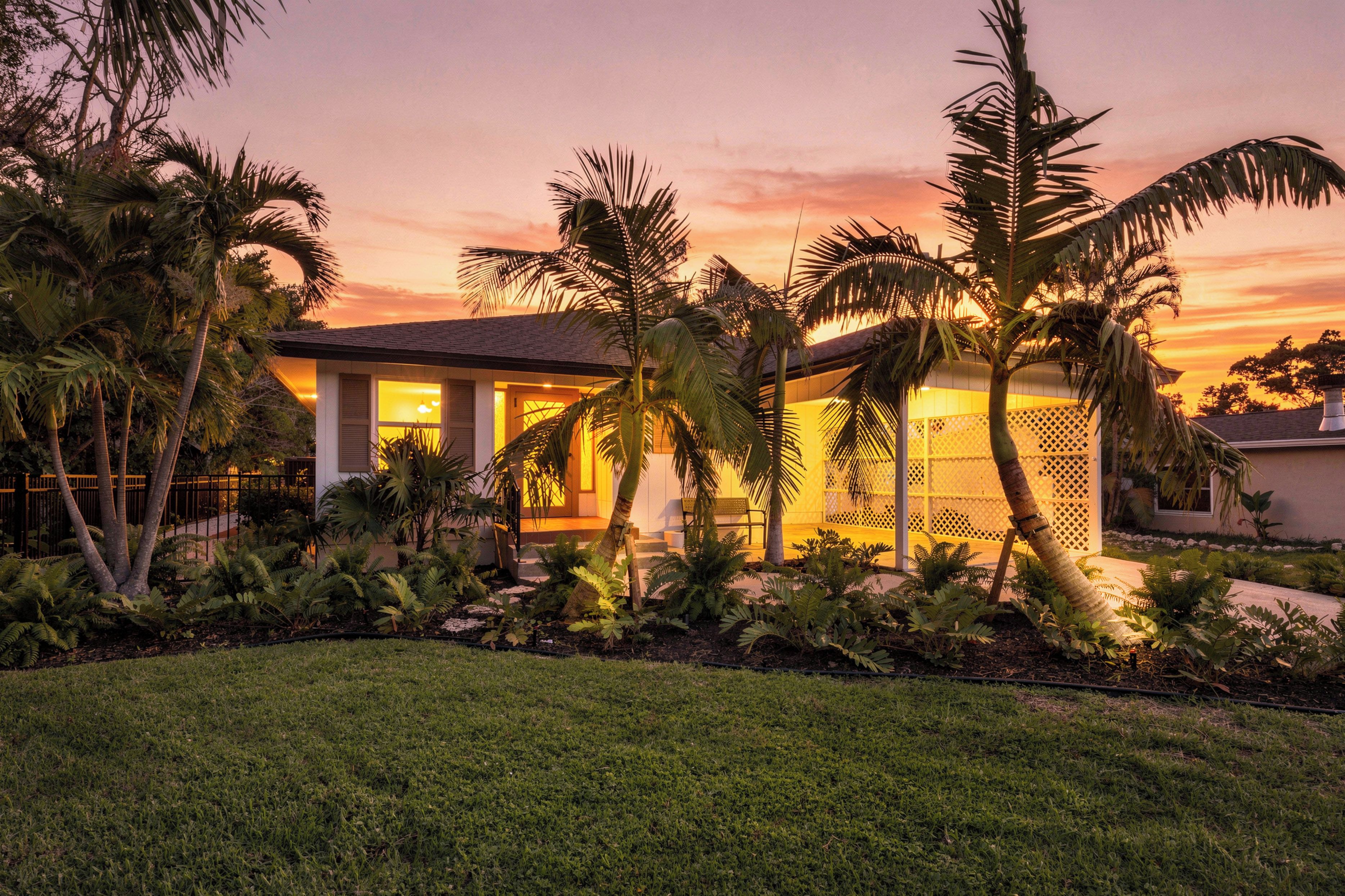 Ein modernes Haus mit einem gepflegten Garten, darunter Palmen und Farne, im Abendlicht.