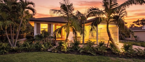 Una casa moderna con un jardín cuidado, que incluye palmeras y helechos, al atardecer.